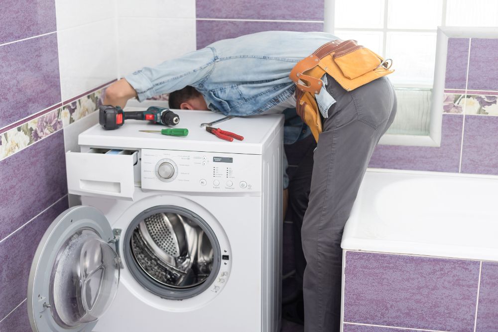 A man working on a washing machine in a bathroom, illustrating laundry appliance repair services available in Washington, DC.