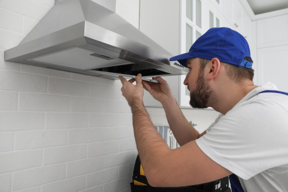 A man in a blue hat and white shirt is fixing a stove hood, highlighting appliance repair services in Washington, DC.