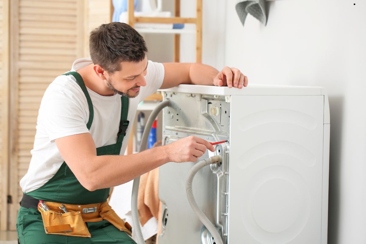 A maintenance worker adjusts a washing machine's connections, tools in a belt, surrounded by household items in a bright room.