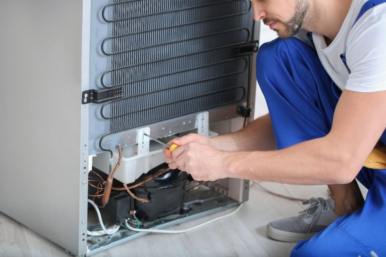 A technician wearing blue overalls works on the back of a refrigerator, using a screwdriver to access internal components.