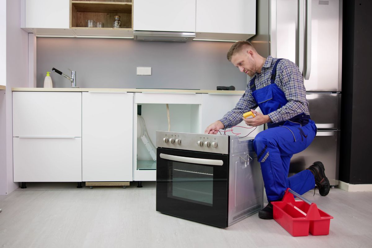 A technician in blue overalls kneels beside a black oven, checking wires with a multimeter in a modern kitchen setting.