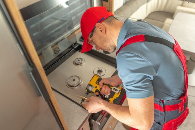 A person in a red cap and overalls is using a drill to install components on a modern kitchen stovetop in an RV.
